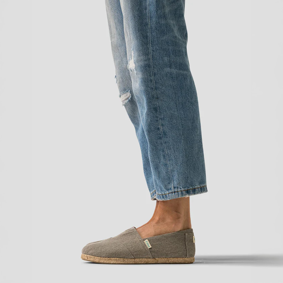 A side profile of a person wearing grey slip-on shoes with cork soles and ripped blue jeans, standing against a plain grey background.