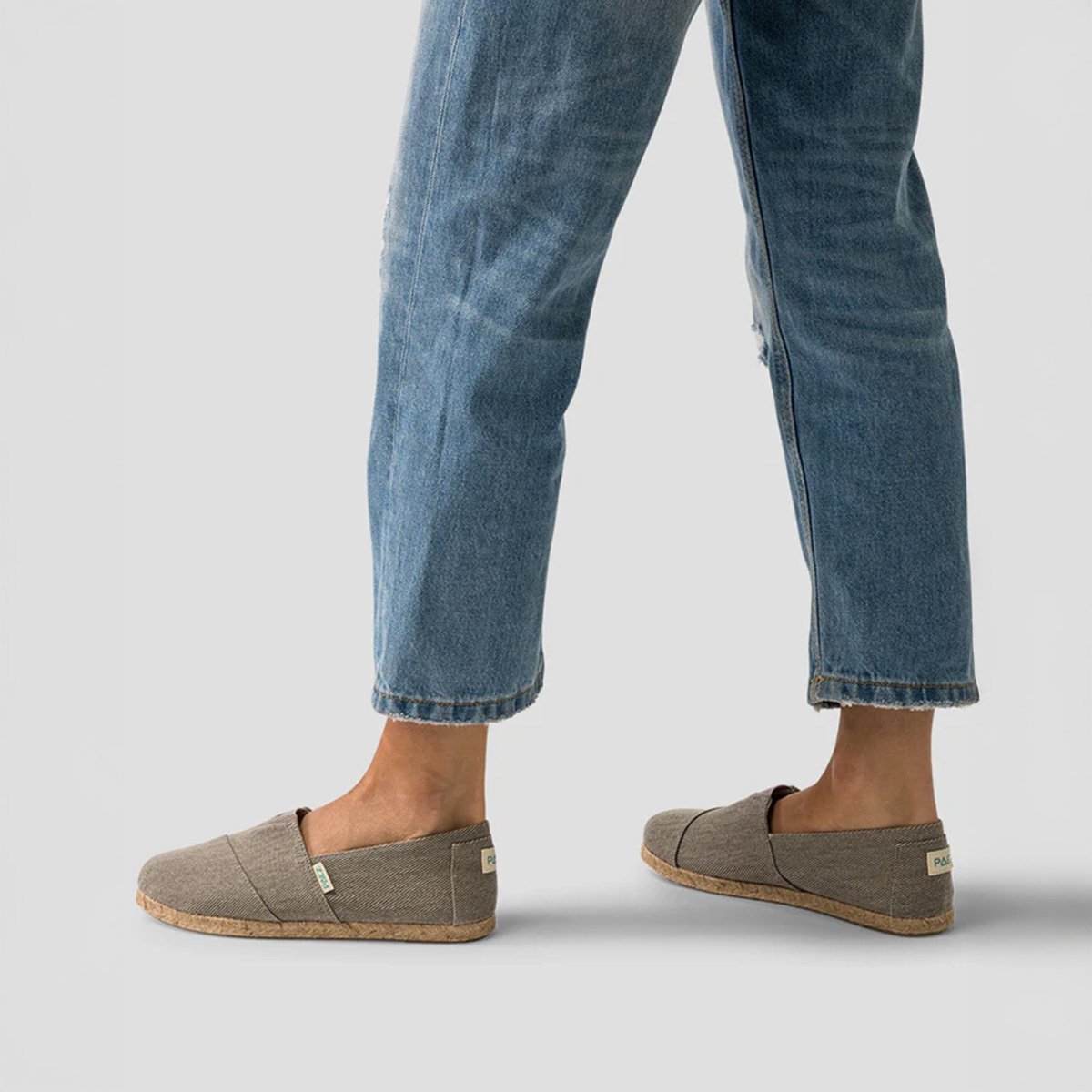 A person wearing rolled-up blue jeans paired with grey slip-on shoes with cork soles, standing against a plain grey background.