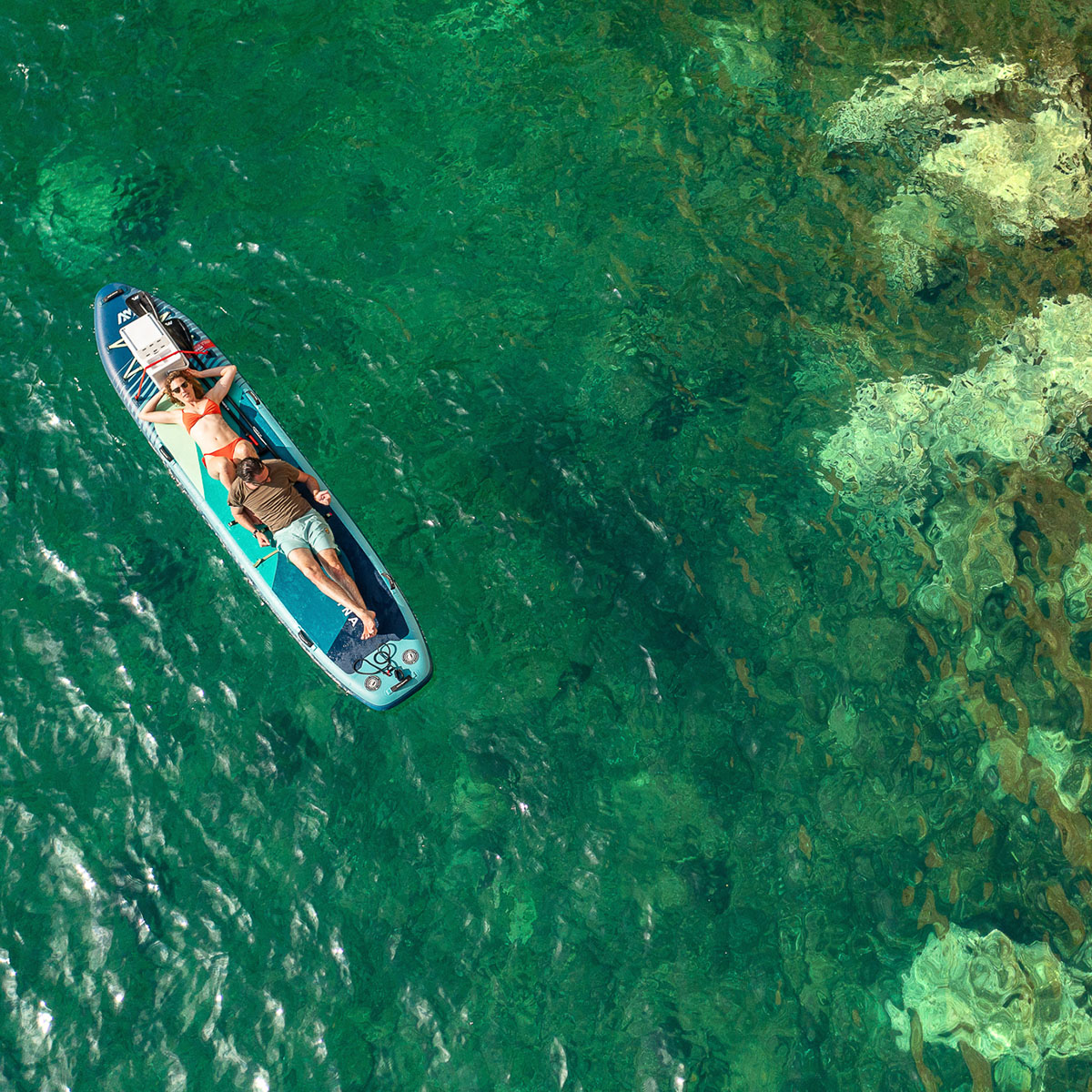 A couple relaxing on an Aqua Marina Super Trip Tandem inflatable SUP floating over crystal-clear water.