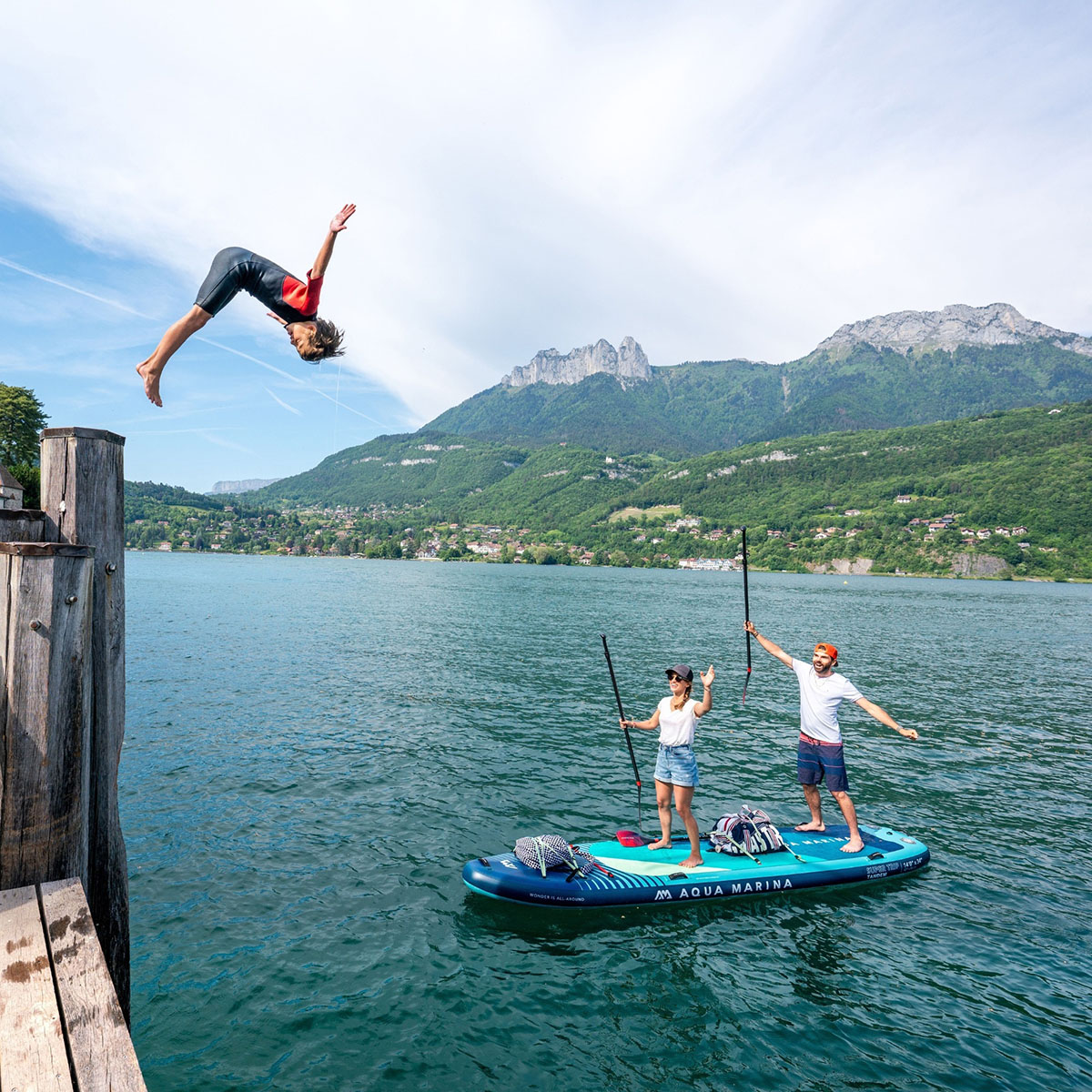 A child performs a backflip off a wooden dock, while two adults on an Aqua Marina SUPER TRIP TANDEM inflatable SUP cheer with their paddles against a scenic mountain backdrop.