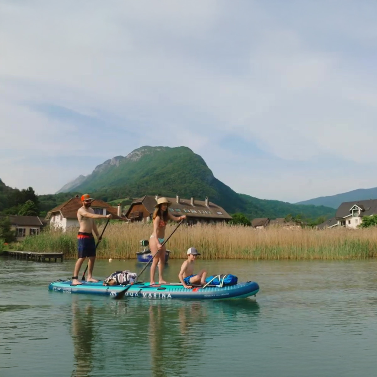 A family of three paddleboarding on an Aqua Marina SUPER TRIP TANDEM inflatable SUP, gliding through a calm river with scenic mountains and countryside houses in the background.