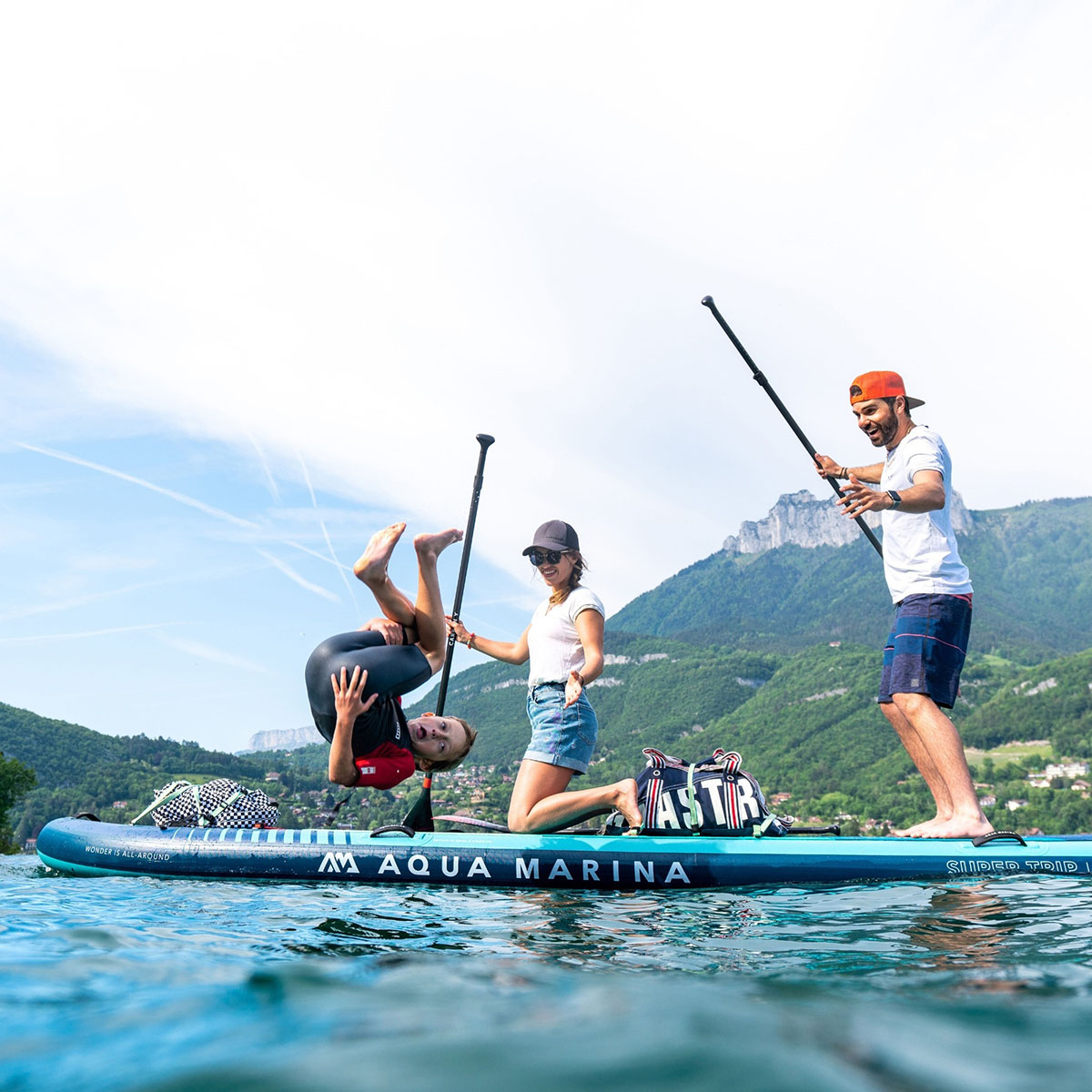 A child in a wetsuit flips off an Aqua Marina SUPER TRIP TANDEM inflatable SUP, while two adults laugh and cheer against a stunning mountain and lake backdrop.