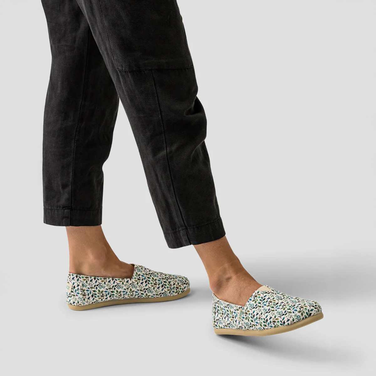 A pair of PAEZ floral emerald espadrilles worn by a person styled with black pants, showing both feet in motion.