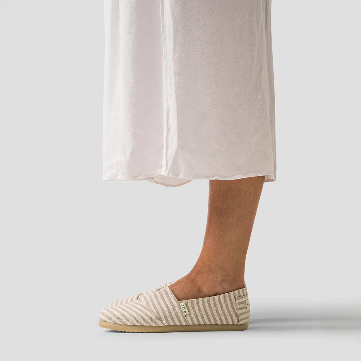 A side view of Sand PAEZ Surfy espadrille shoes for women, featuring beige and cream striped fabric, worn with a flowy white dress against a plain grey background.